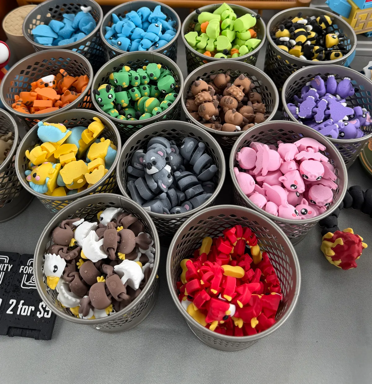 Rows of round mesh baskets on the stall table, each filled with one design of 3D printed articulated mini animal, including koalas, bees and eagles