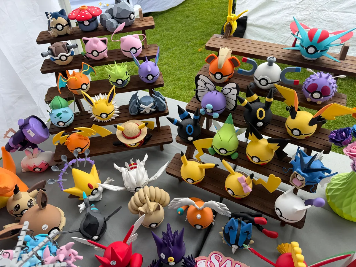 Two tiered wooden shelves of 3D printed Pokeball figures in many colours on the Formfinity market stall, with grass and a white marquee behind