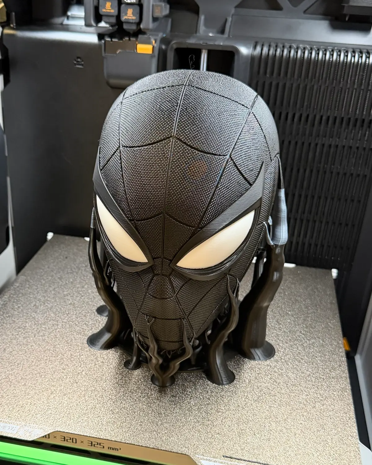 A black Spider-Man head headphone stand with white eyes and a woven suit texture, on the build plate of a Bambu Lab H2D 3D printer with branching tree supports still attached